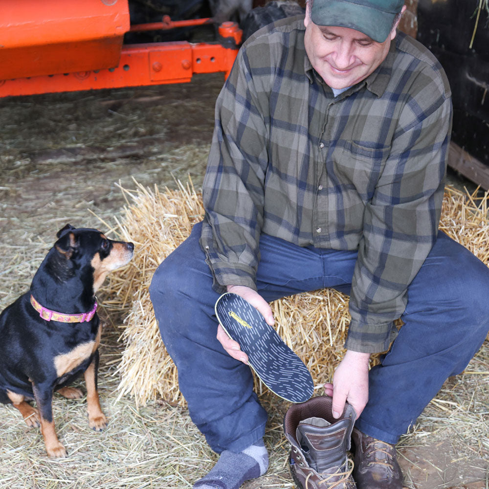 Man sitting on bale of hay next to his dog, placing PowerStep Pinnacle Work insole into work boot before working on the farm