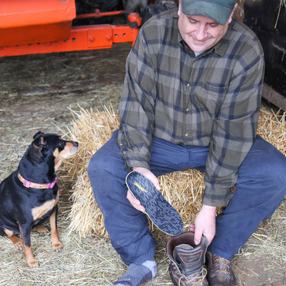 Man sitting on bale of hay next to his dog, placing PowerStep Pinnacle Work insole into work boot before working on the farm