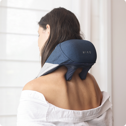 A woman seen from behind wearing a white off-the-shoulder shirt, using a navy blue neck and shoulder massager with the brand name 'MIKO' visible on the device. The background is a bright, softly lit room with white window blinds.